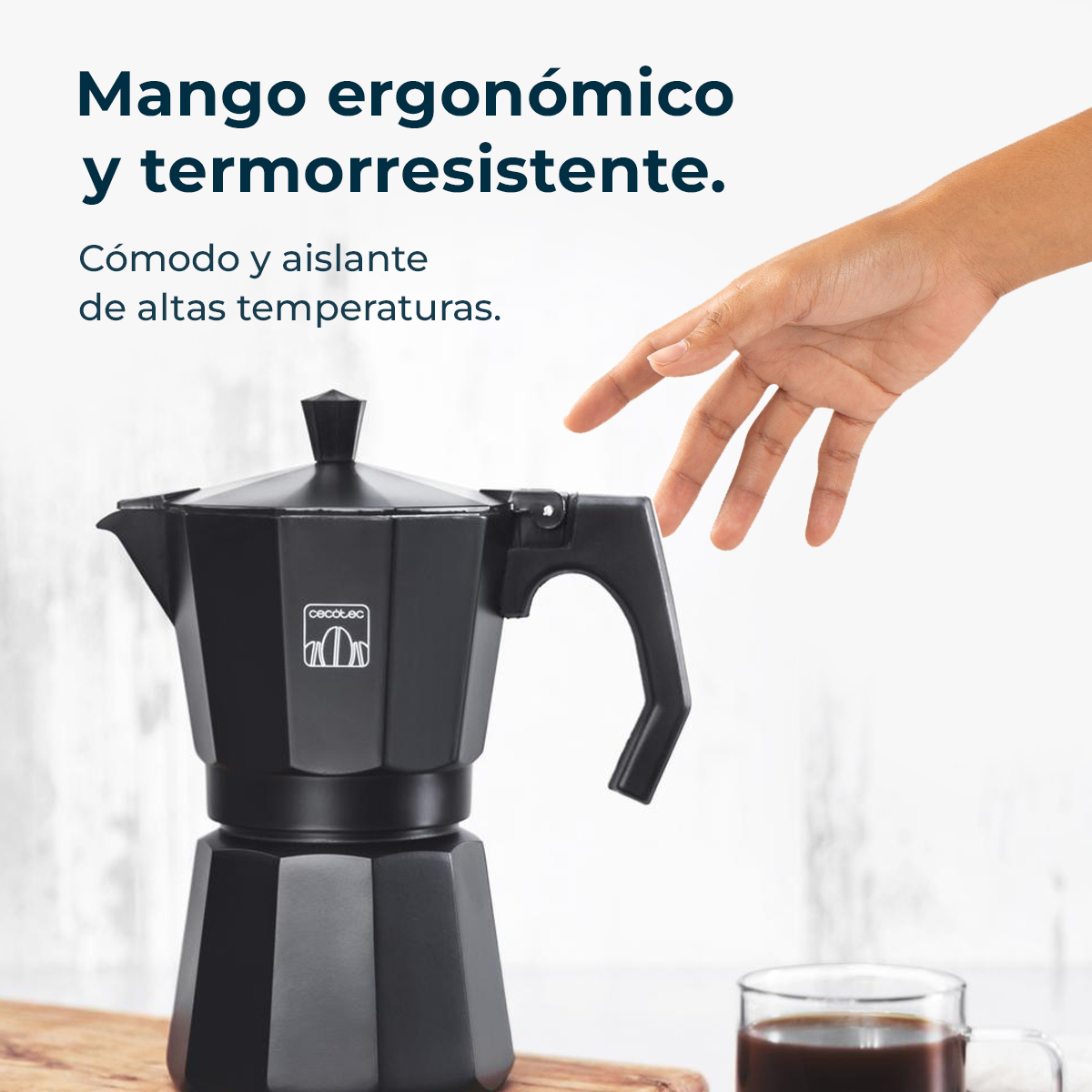 Una cafetera moka negra con una mano extendiéndose hacia el asa. Una taza de café transparente abajo a la derecha.