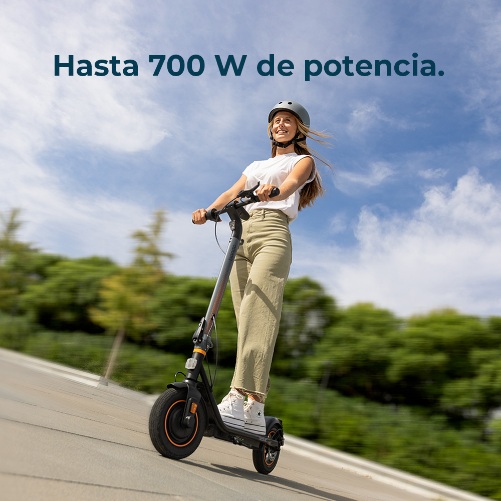 Una donna guida un monopattino elettrico. Indossa un casco e sorride. Il cielo è blu con nuvole. Il testo recita: Hasta 700 W de potencia.