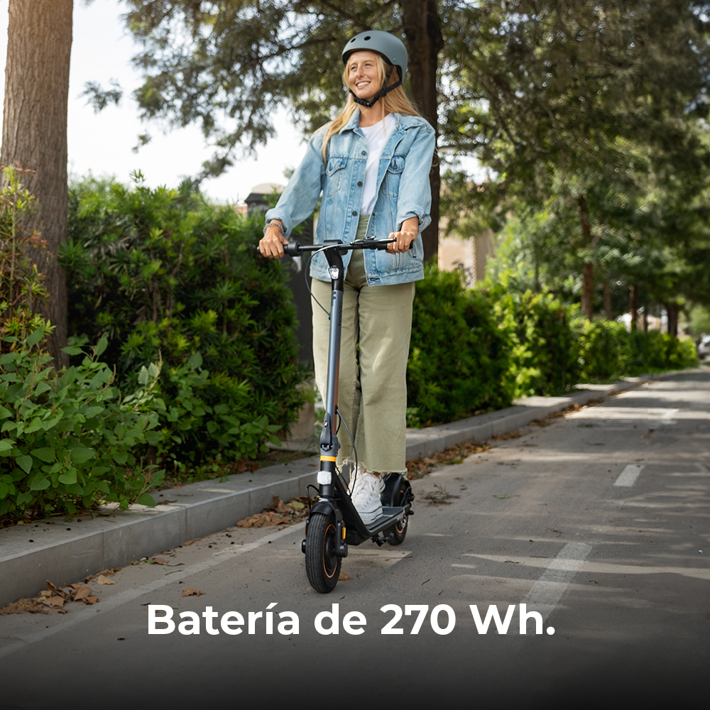 Una donna guida uno scooter su una strada. Indossa un casco e sorride. Alberi e cespugli sullo sfondo. Il testo recita 'Batería de 270 Wh.'