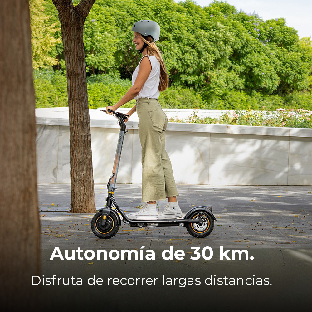 Una donna con un casco guida un monopattino elettrico. È in piedi vicino a un albero. Fogliame verde sullo sfondo.