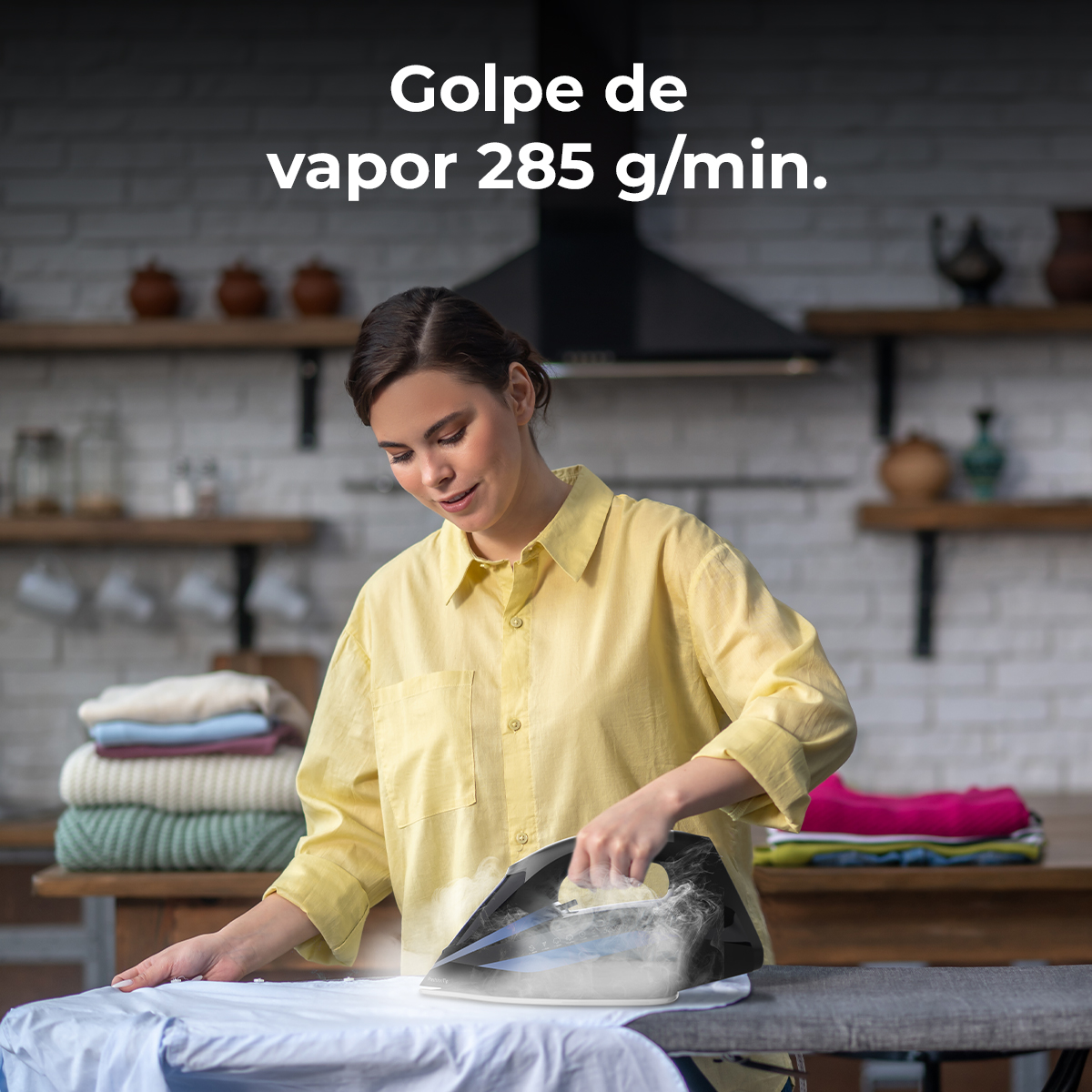 Eine Frau bügelt ein weißes Hemd. Dampf kommt aus dem Bügeleisen. Kleidung ist auf dem Tisch gestapelt. Der Text „Golpe de vapor 285 g/min“ ist sichtbar.