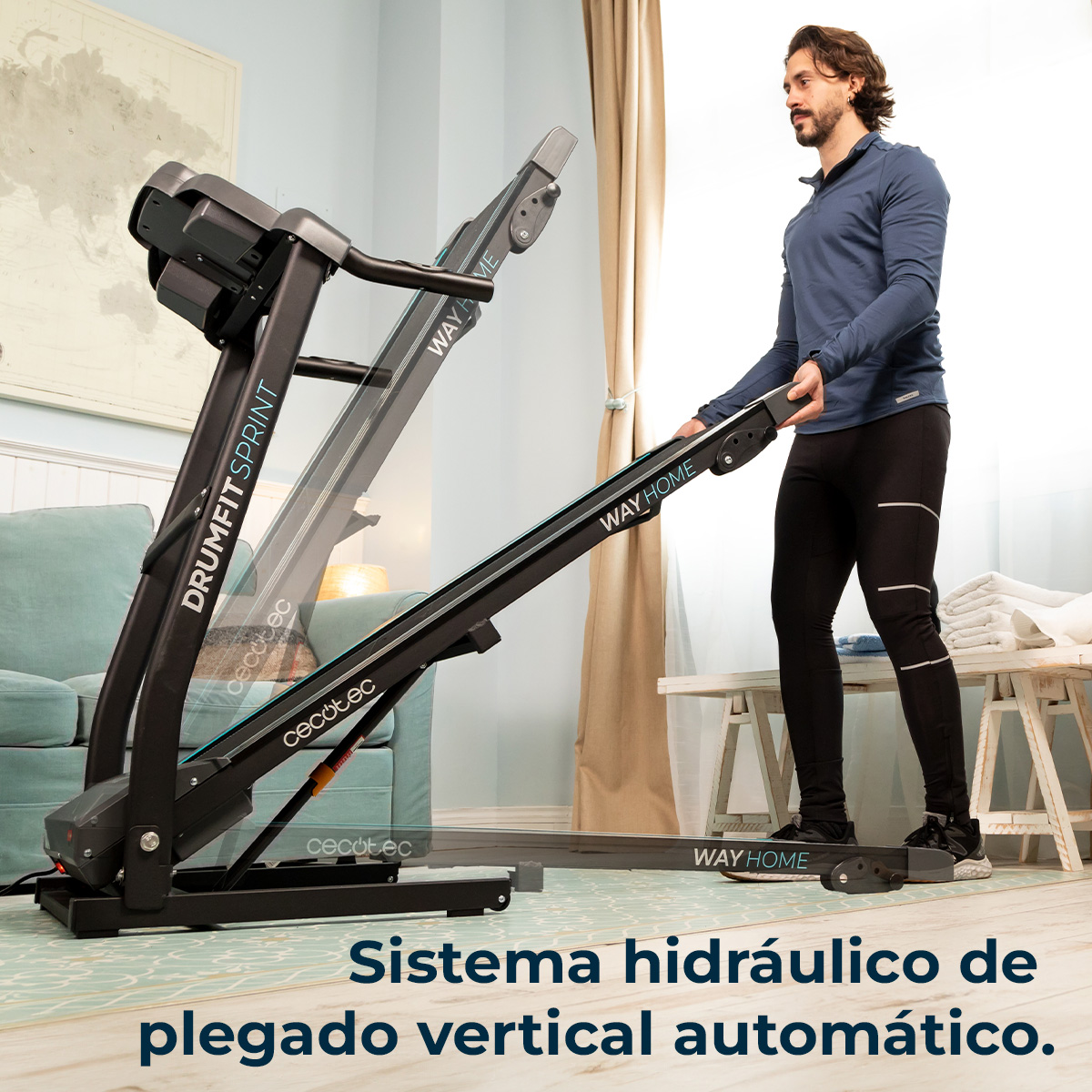 Mężczyzna składa bieżnię. Bieżnia jest czarna z białymi etykietami. W tle kanapa, stół i mapa.