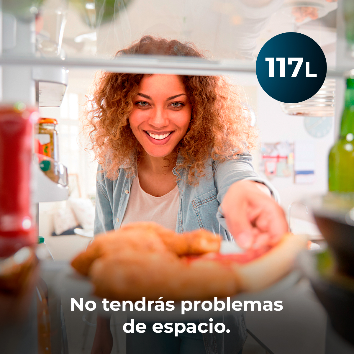 Una mujer sonríe, metiendo la mano en una nevera. Pizza visible. Adhesivo 117L al fondo. Texto: No tendrás problemas de espacio.
