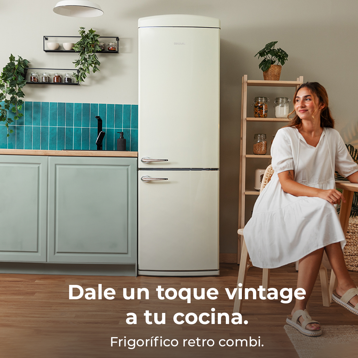 Kobieta w białej sukience siedzi obok lodówki retro. Kuchnia ma turkusowe kafelki i drewniane półki z roślinami.