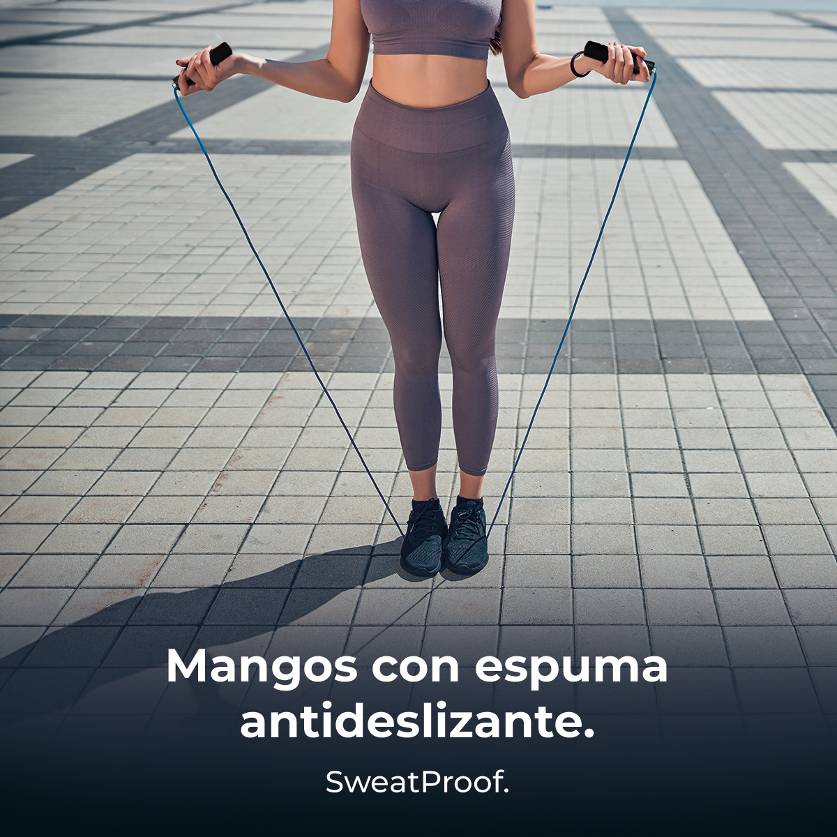 Una donna salta la corda all'aperto. Indossa abiti da allenamento grigi e scarpe da ginnastica nere. La corda è blu, il pavimento è piastrellato.