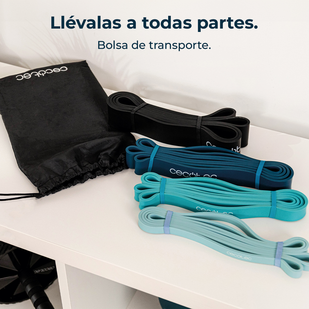 Bolsa negra y bandas de resistencia en negro, verde azulado y aguamarina sobre una superficie blanca. El texto dice: Llévalas a todas partes. Bolsa de transporte.