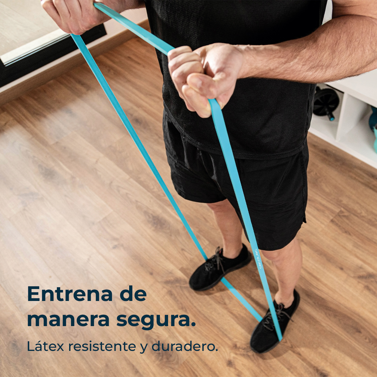 Una persona se ejercita con una banda de resistencia azul. Viste pantalones cortos y zapatos negros, en un piso de madera.