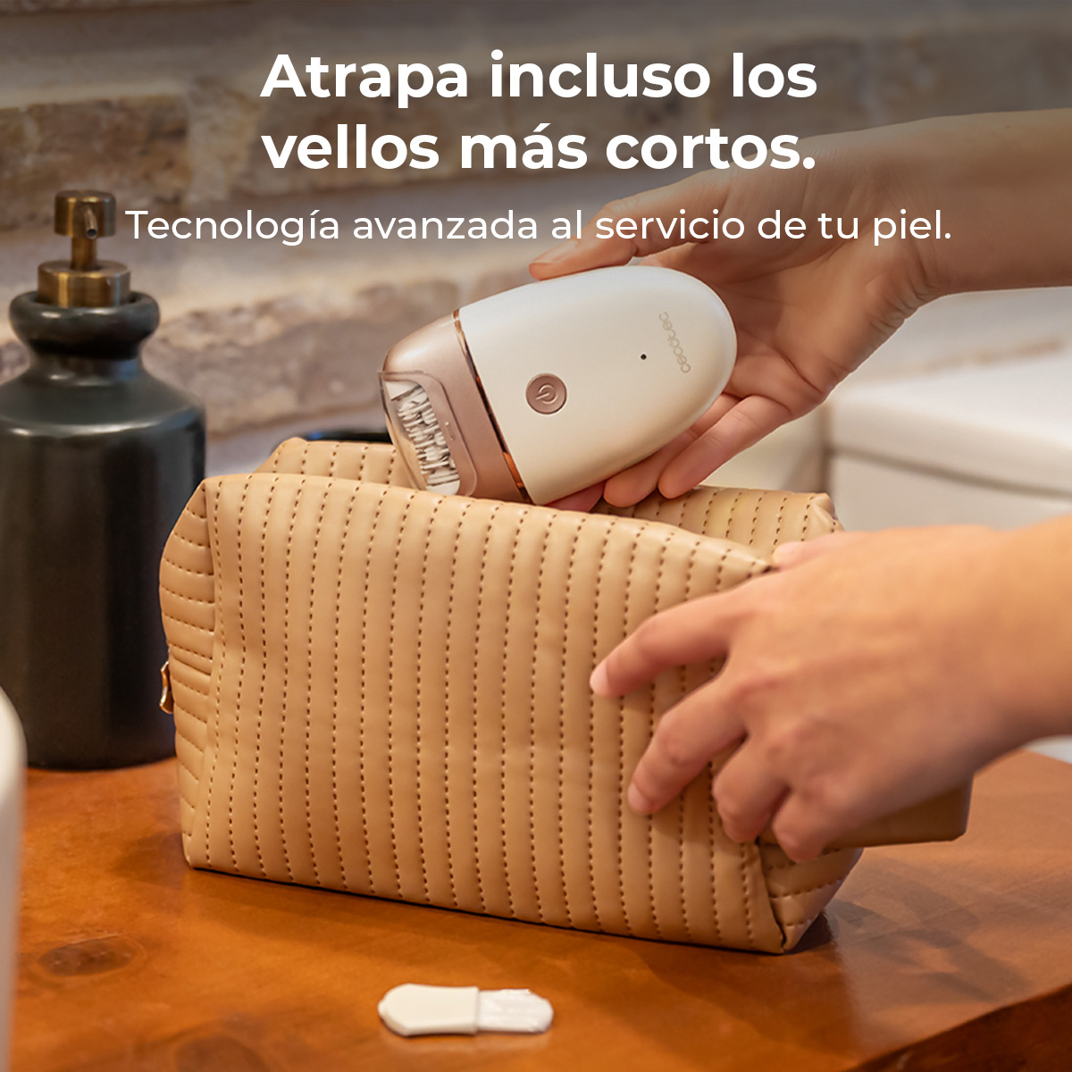 Manos sostienen una depiladora blanca y dorada sobre un neceser beige. Una botella negra a la izquierda, un cepillo abajo.