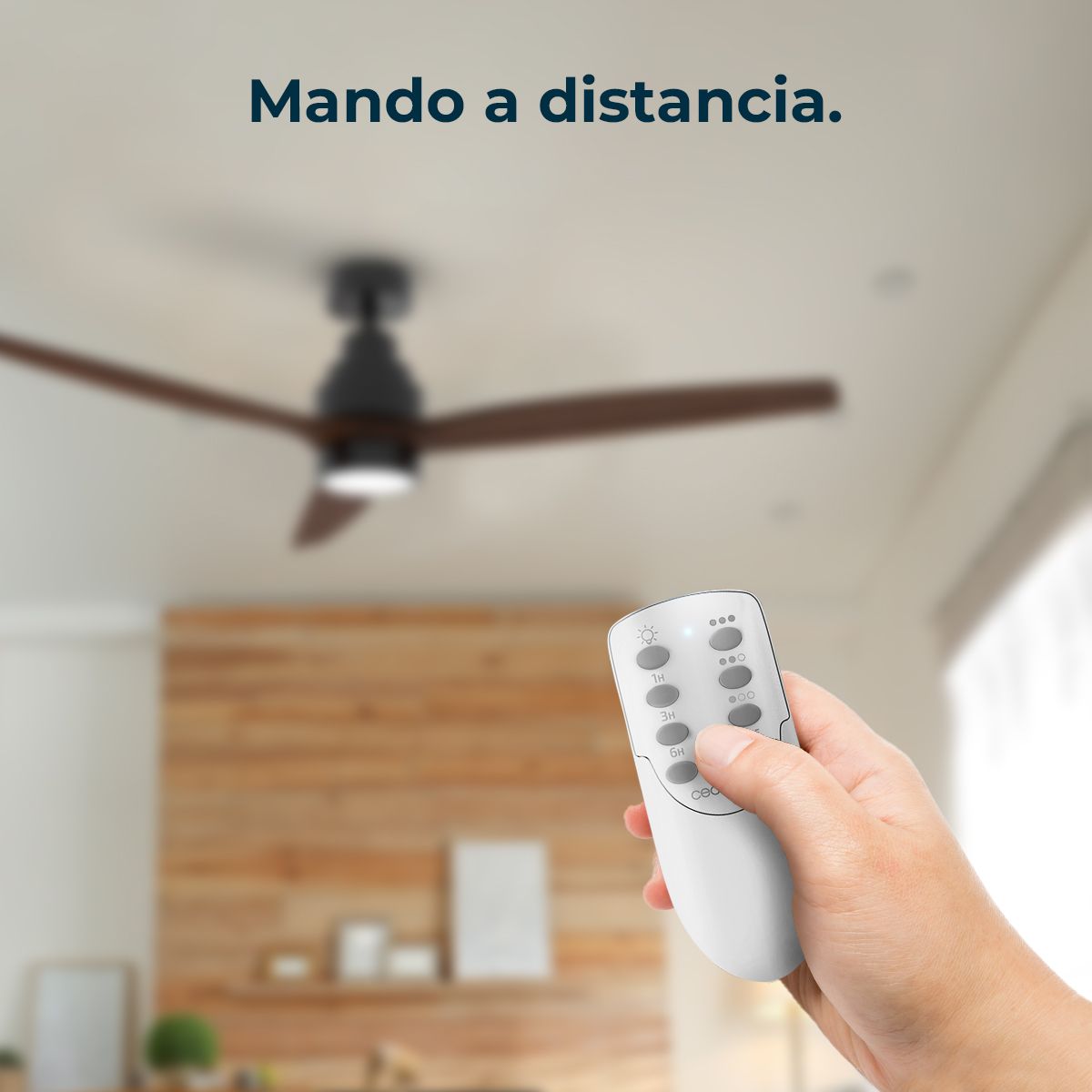 Una mano sostiene un control remoto blanco, controlando un ventilador de techo. El ventilador tiene aspas oscuras. El fondo muestra una pared de madera.