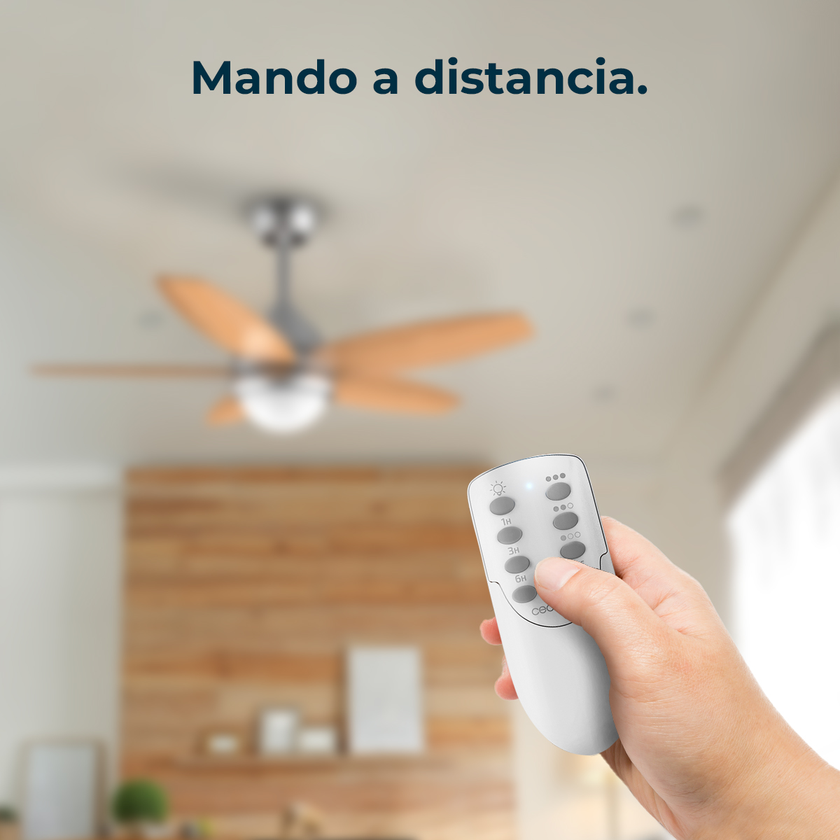 Dłoń trzyma biały pilot. Rozmyty wentylator sufitowy w tle. Tekst 'Mando a distancia' na górze.