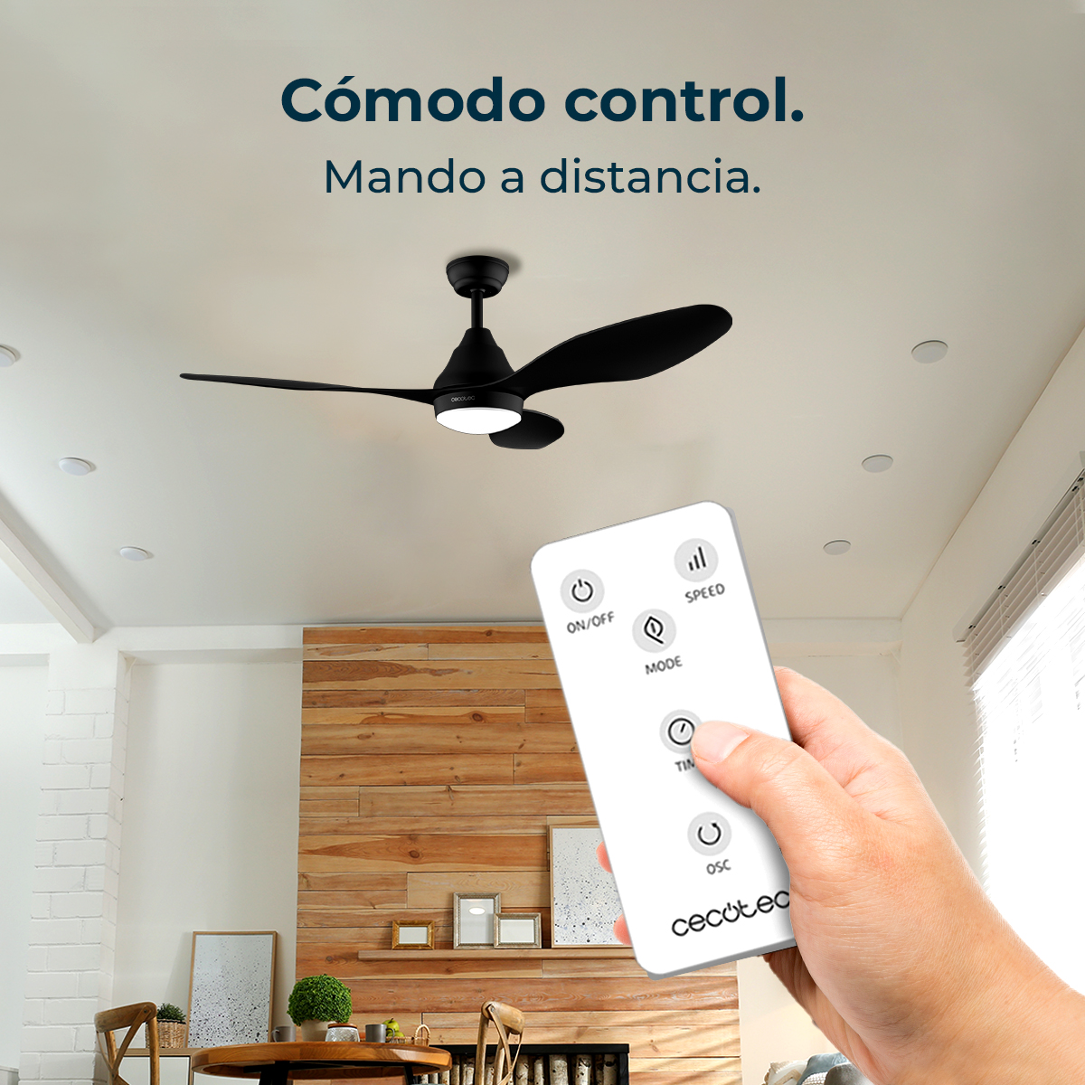 Ręka trzyma pilota. Czarny wentylator sufitowy jest u góry. Drewniana ściana w tle. Tekst: Comodo control, Mando a distancia.