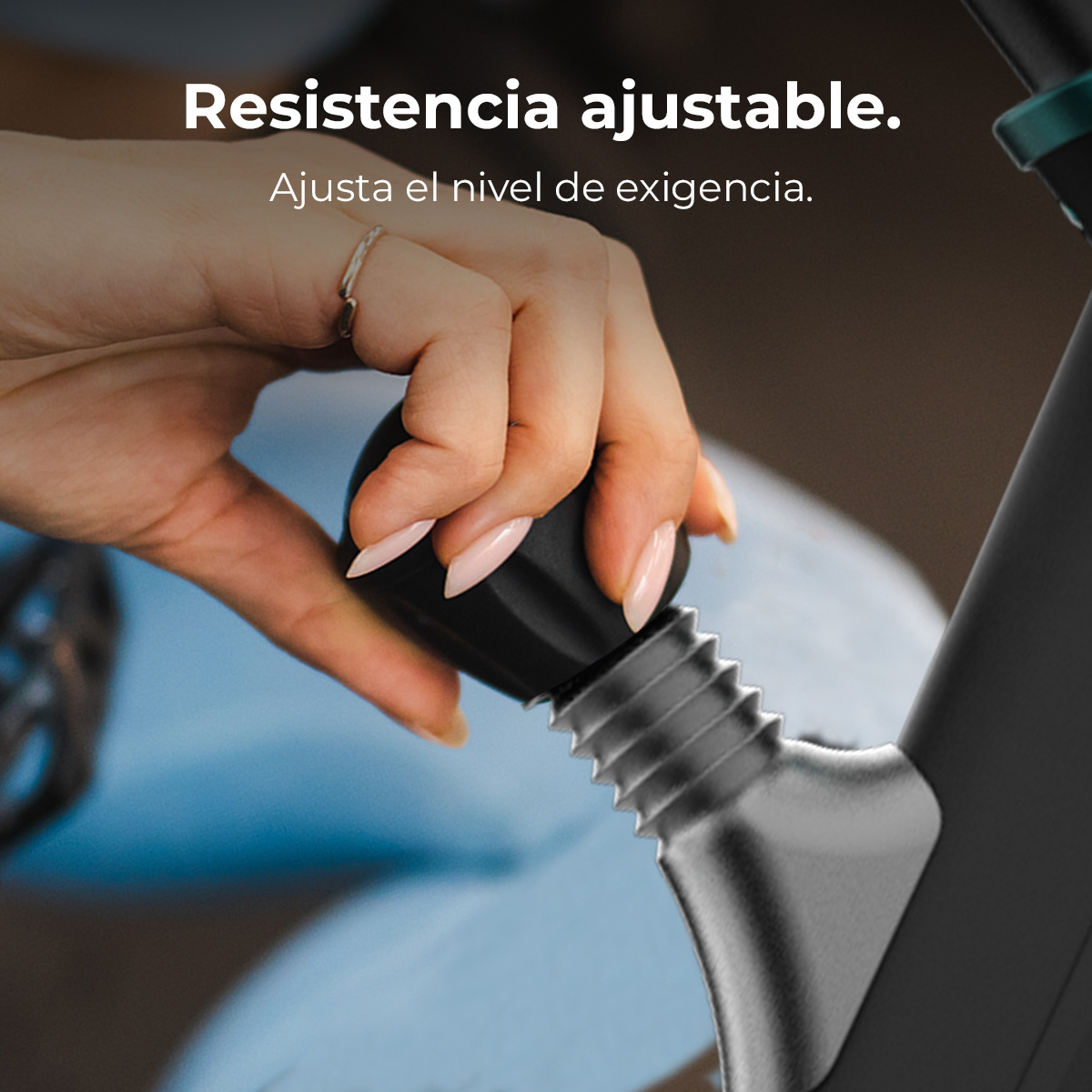 Ręka reguluje pokrętło oporu w rowerze stacjonarnym. Pokrętło jest czarne, dłoń ma pierścionek i jasnoróżowe paznokcie.