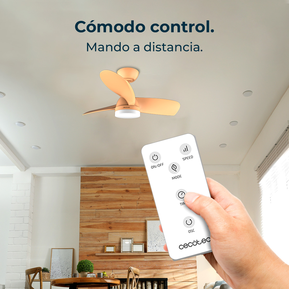 Una mano sostiene un mando a distancia blanco. Arriba hay un ventilador de techo. Una pared de madera y un salón en el fondo.