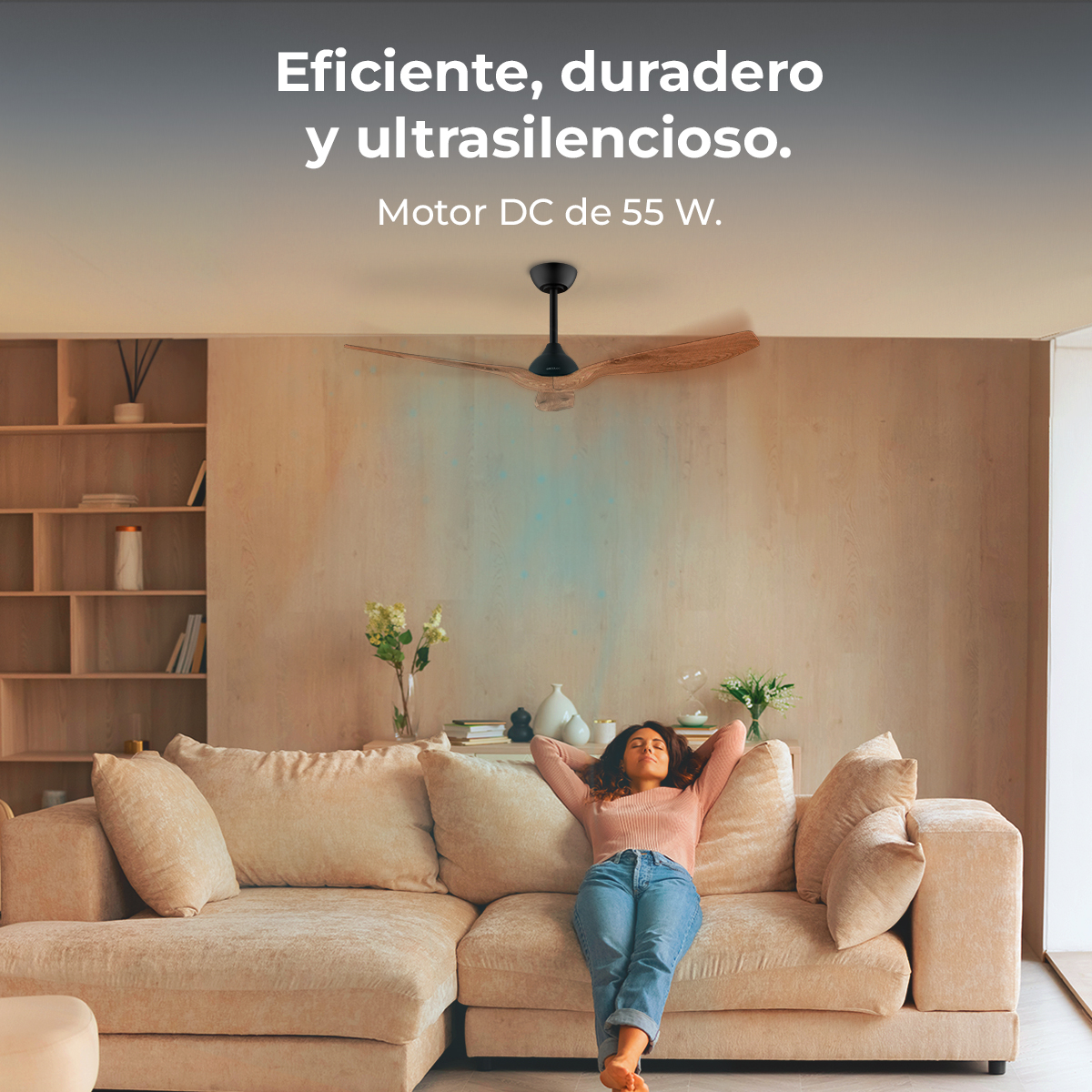 Una mujer se relaja en un sofá beige, con los brazos detrás de la cabeza. Un ventilador de techo está arriba. La habitación tiene paredes claras y una estantería.
