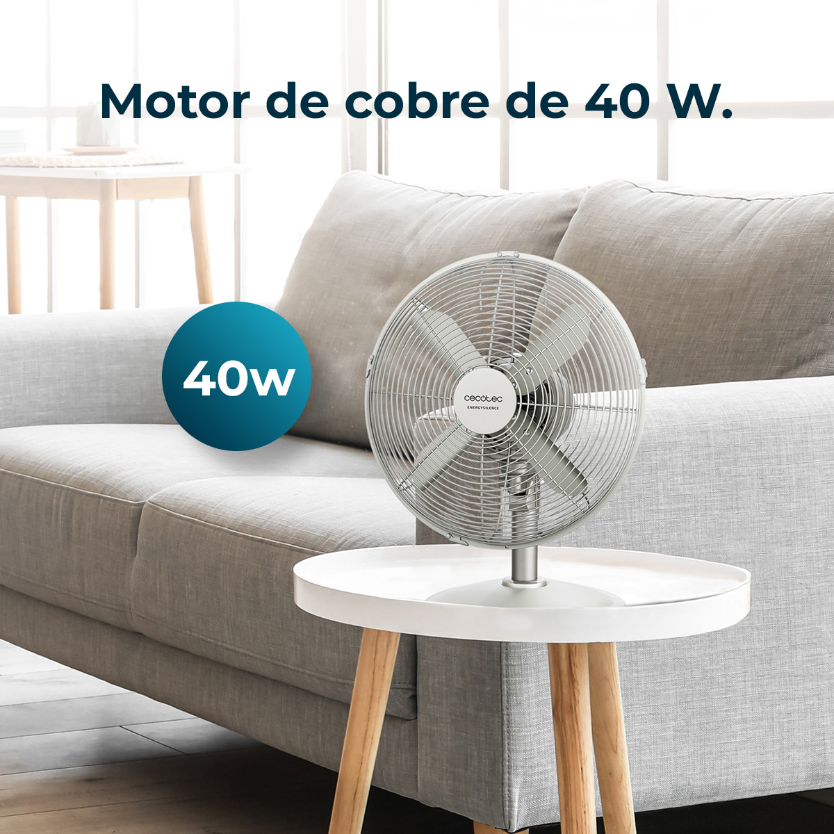 Metalowy wentylator stoi na białym stole. Za nim szara sofa. Niebieskie koło pokazuje moc 40W. Tekst: 'Motor de cobre de 40 W'.