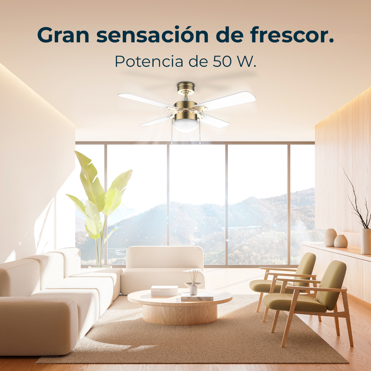 Un salón moderno con ventilador de techo, sofá, sillones y mesa de centro redonda. Grandes ventanales muestran vistas a la montaña.