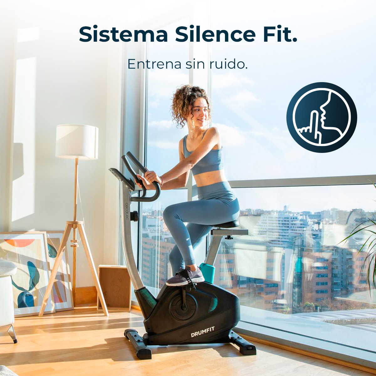 Donna su cyclette indoor. La bici è nera. Indossa abbigliamento sportivo blu. Testo: Sistema Silence Fit. Entrena sin ruido.