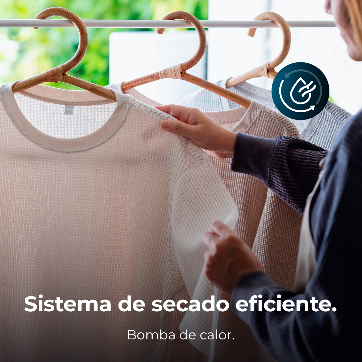 Una persona toca una camisa beige en una percha. Hay otras camisas colgadas cerca. El fondo presenta una ventana con follaje verde.