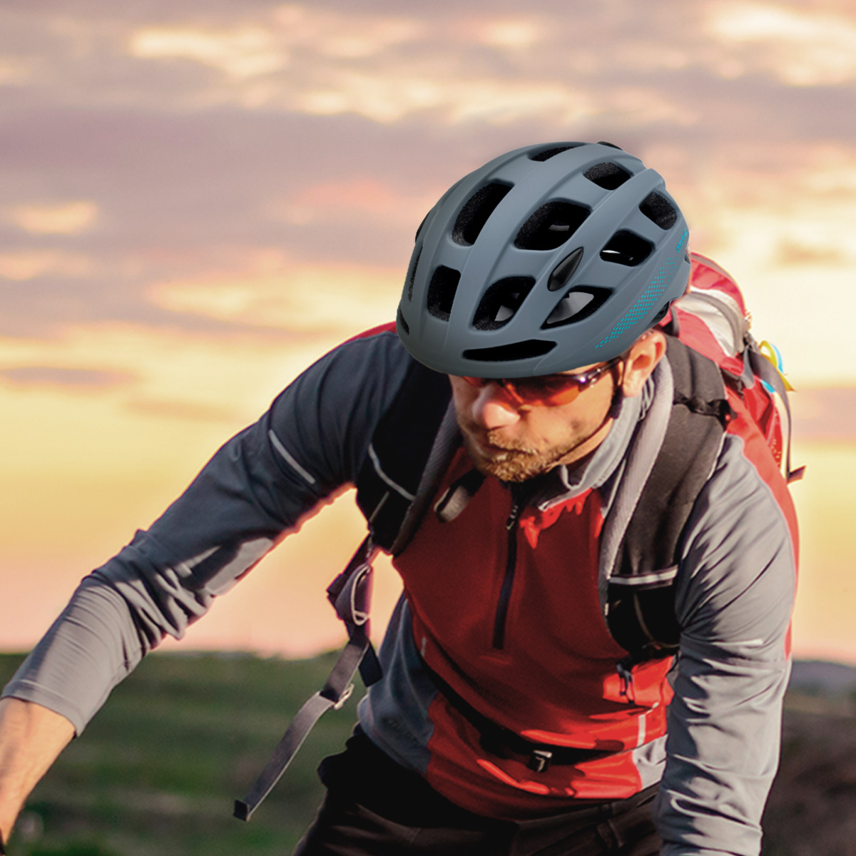 Ciclista con casco gris y gafas de sol, mochila roja. El fondo muestra una puesta de sol naranja y amarilla.