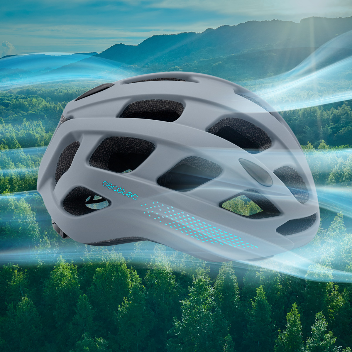 Un casco de bicicleta gris con detalles azules, contra un fondo de bosque verde y montañas bajo un cielo azul.