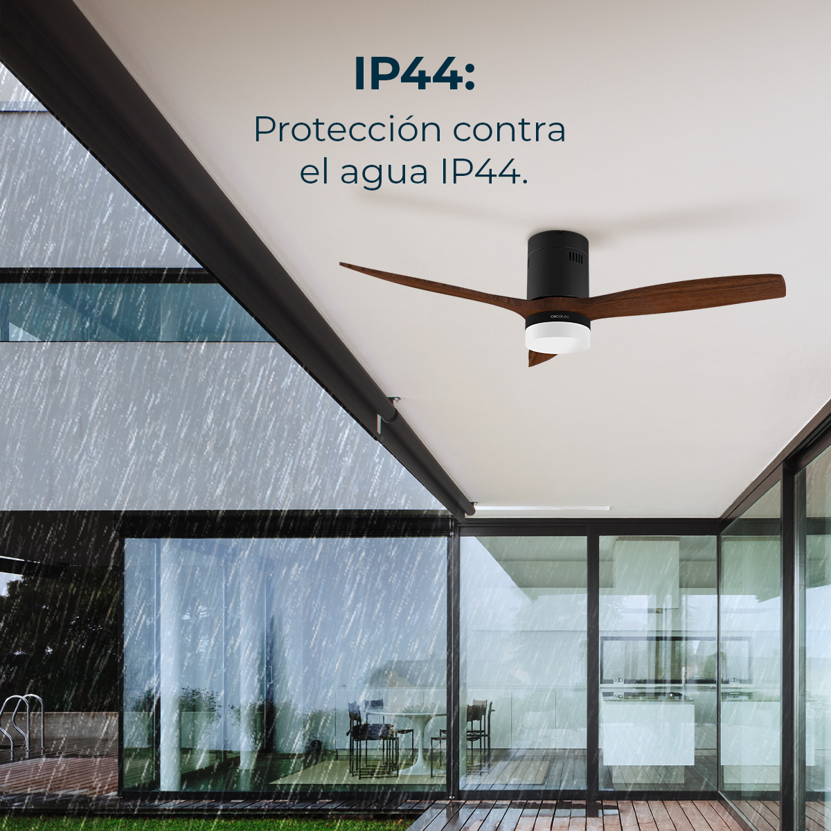 Un ventilador de techo con aspas de madera está instalado en un porche. Llueve. Se ven paredes de vidrio y una cocina moderna en el fondo.