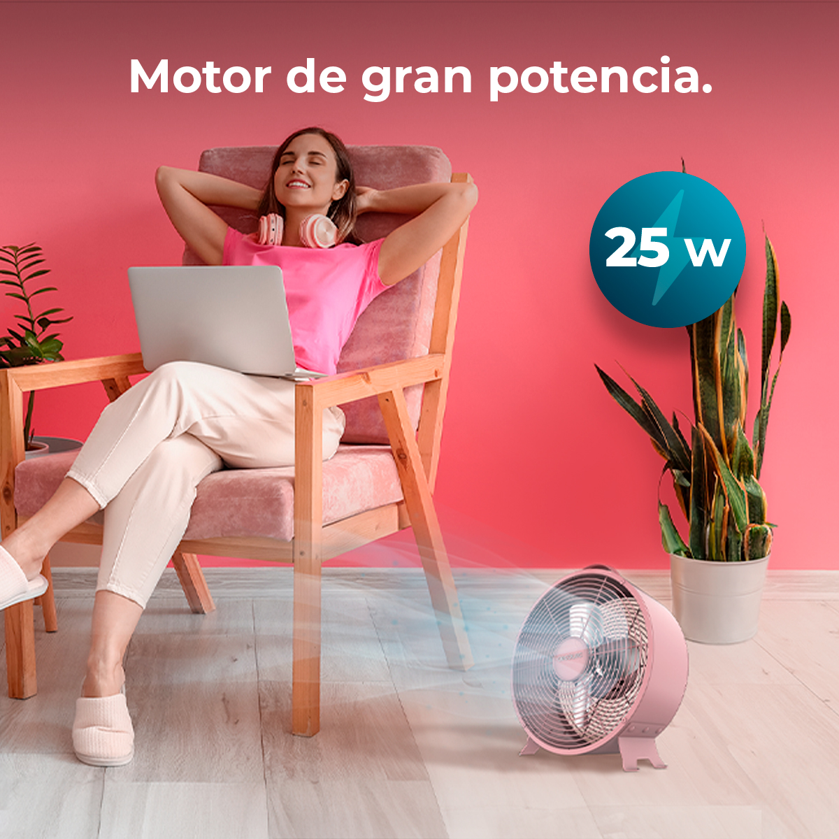 Mujer en rosa relajándose con portátil, ventilador soplando aire. Ventilador y pared rosas, silla de madera, planta e indicador 25W.