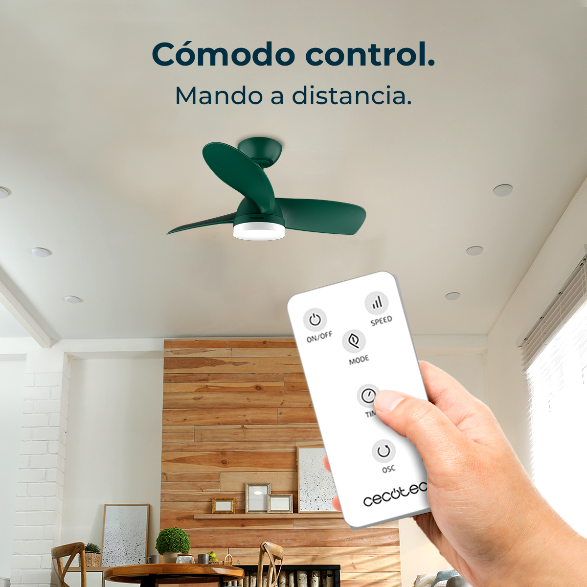Una mano sostiene un mando a distancia que controla un ventilador de techo verde. La habitación tiene una pared de madera.