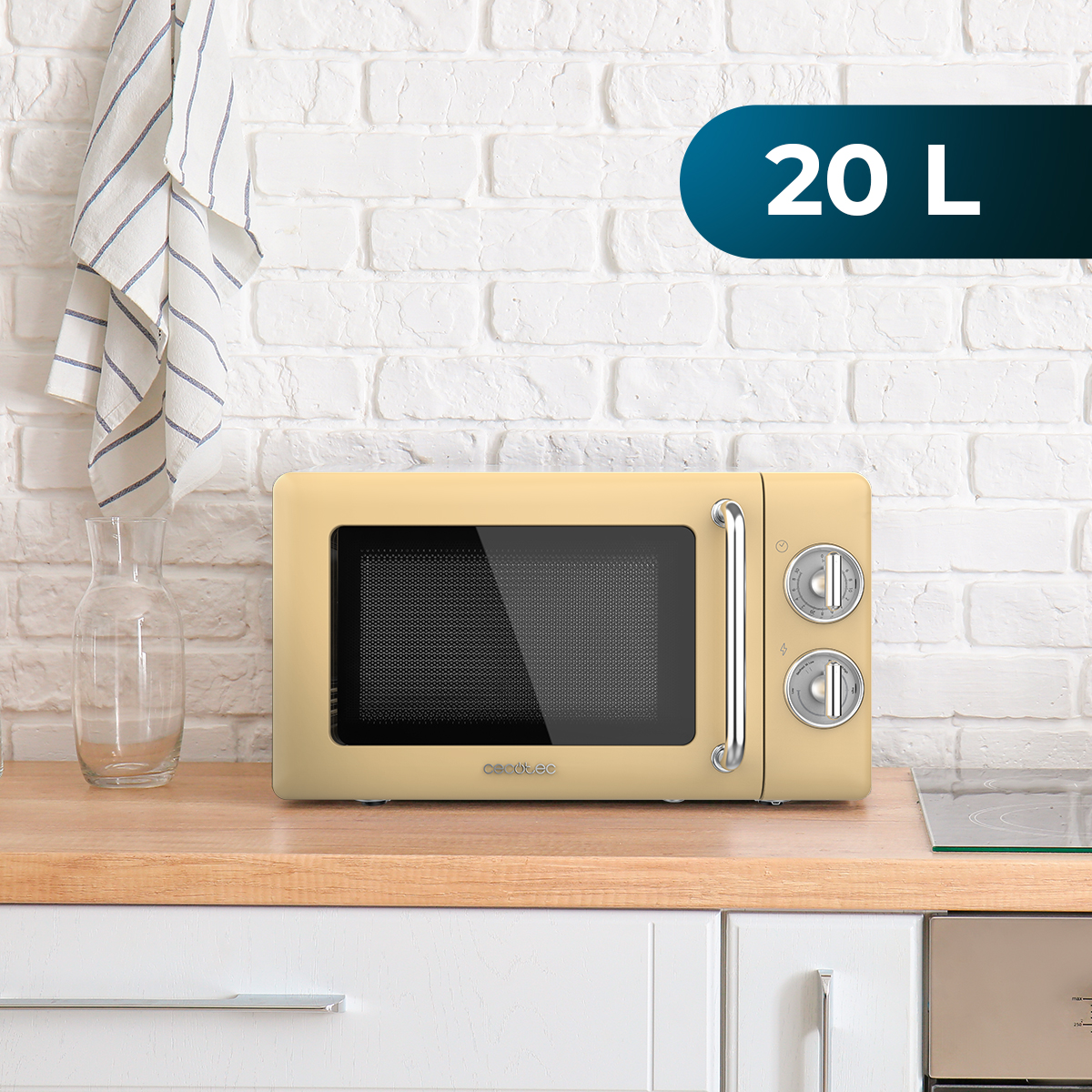 Beżowa kuchenka mikrofalowa na drewnianym blacie. Biała ceglana ściana w tle. Szklany dzbanek i ręcznik po lewej.