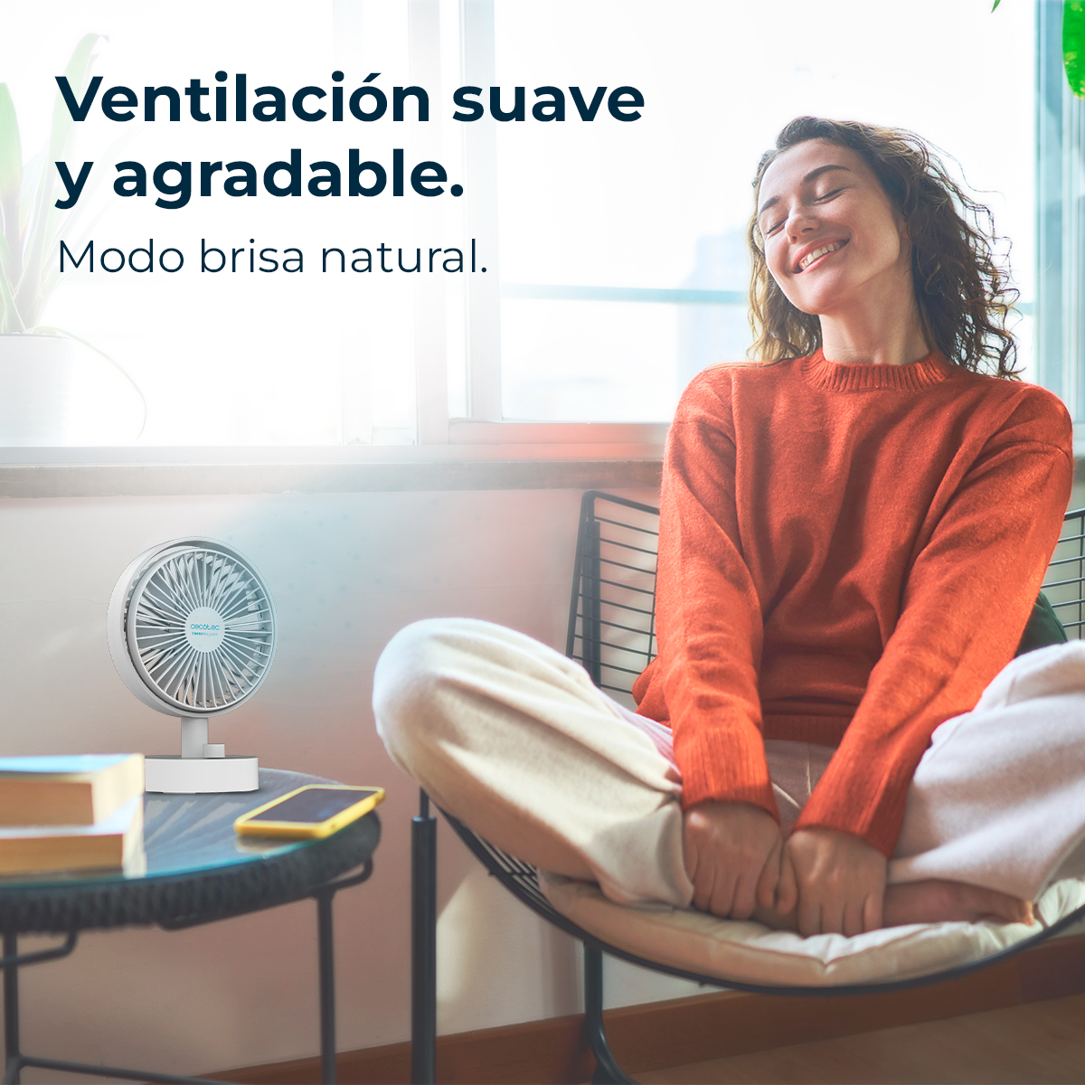 Una donna sorride con gli occhi chiusi, seduta vicino a un ventilatore bianco. Indossa un maglione arancione. Un telefono e dei libri sono su un tavolo di vetro.