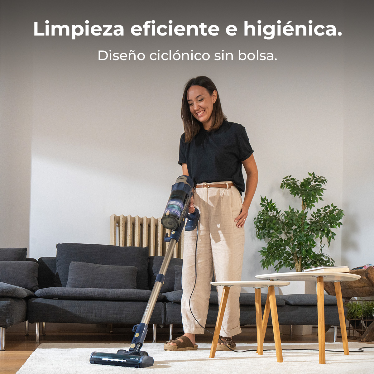 Una mujer aspira una sala de estar con una aspiradora inalámbrica. Hay un sofá, mesas auxiliares y una planta en el fondo.