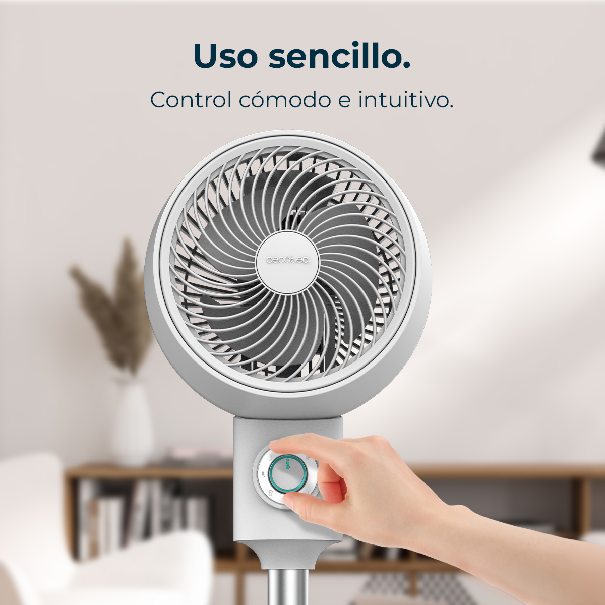 Una mano regola un ventilatore bianco e grigio. Il ventilatore ha una testa circolare e un quadrante di controllo. Sfondo: soggiorno sfocato.