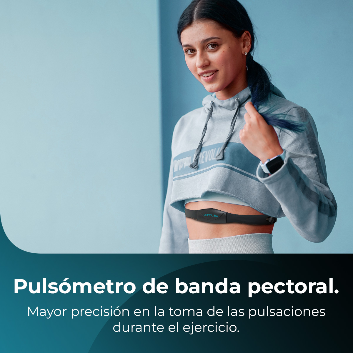 Una donna indossa un cardiofrequenzimetro. Sorride, indossa una felpa con cappuccio corta grigia e uno smartwatch. Un muro blu è dietro di lei.