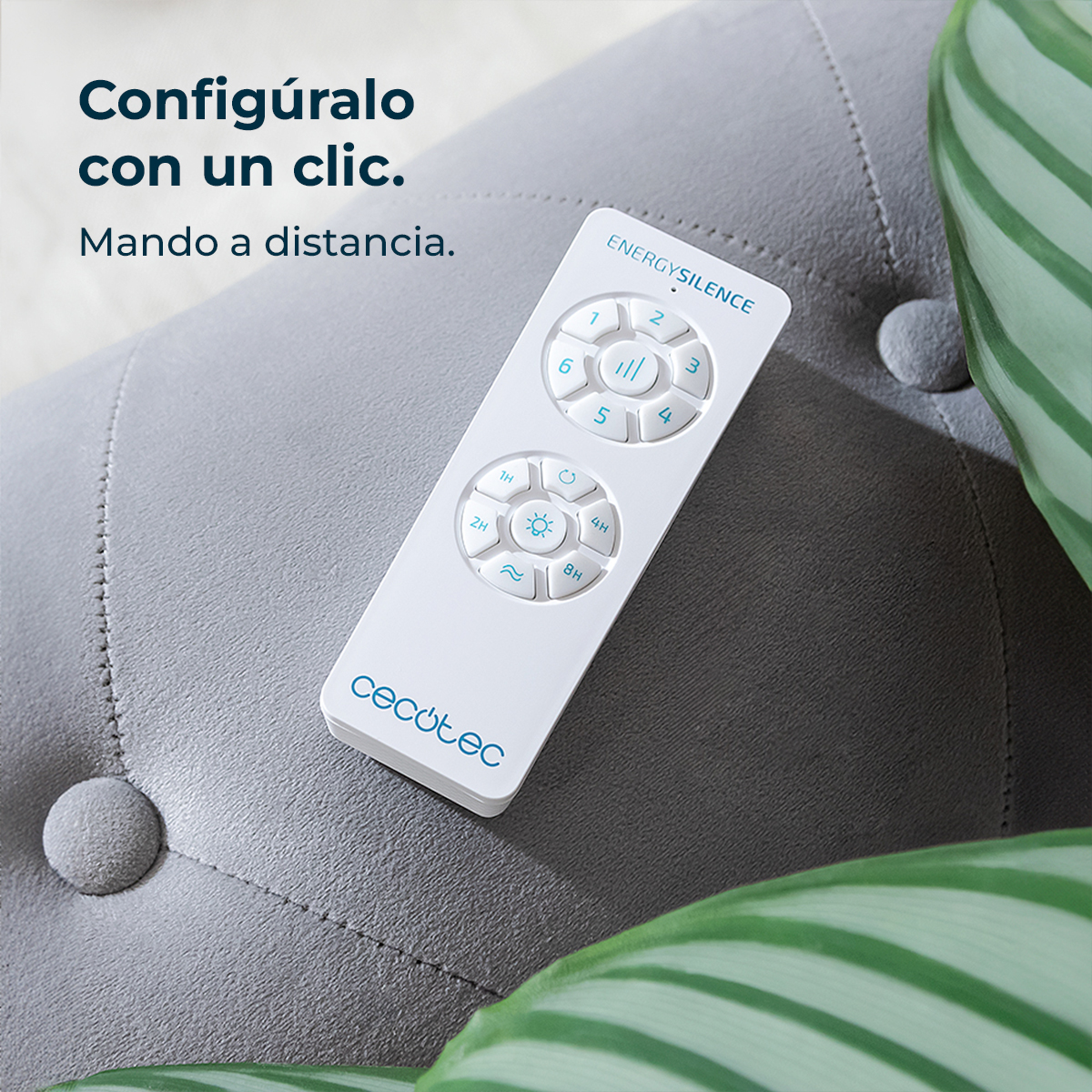 A white remote control lies on a gray cushioned surface. Plant leaves are in the background. The remote has buttons and text on it.