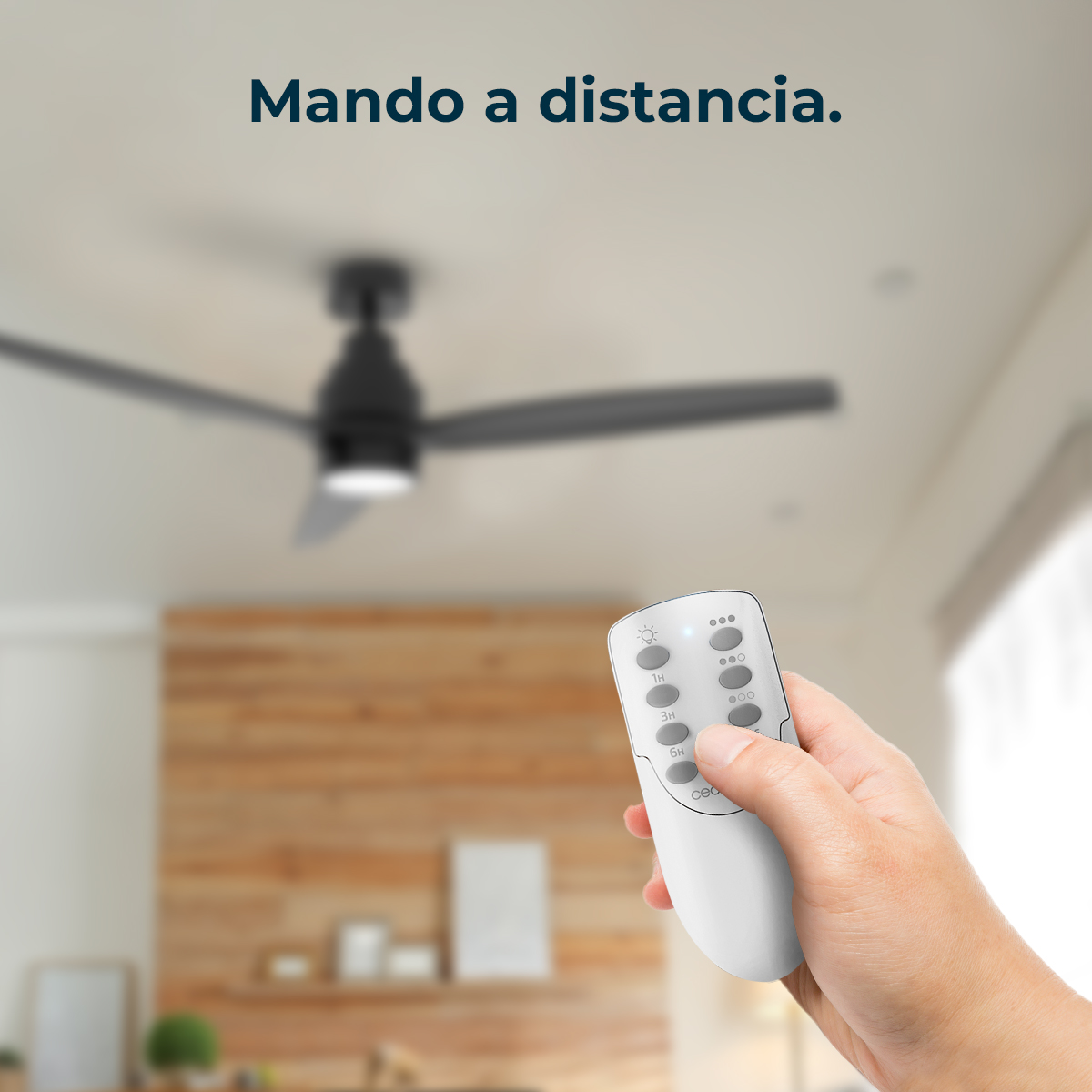Ręka trzyma biały pilot z przyciskami. Powyżej widoczny czarny wentylator sufitowy. Tło przedstawia drewnianą ścianę.