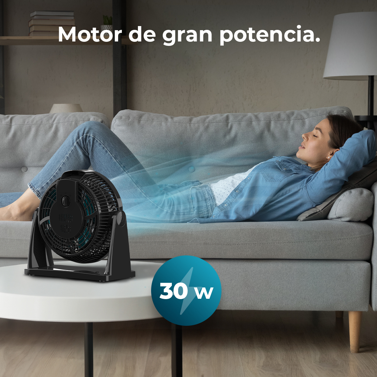 Una donna si rilassa su un divano con un ventilatore che soffia aria. È visibile un'etichetta da 30w. La scena ha un tavolo bianco e una lampada.