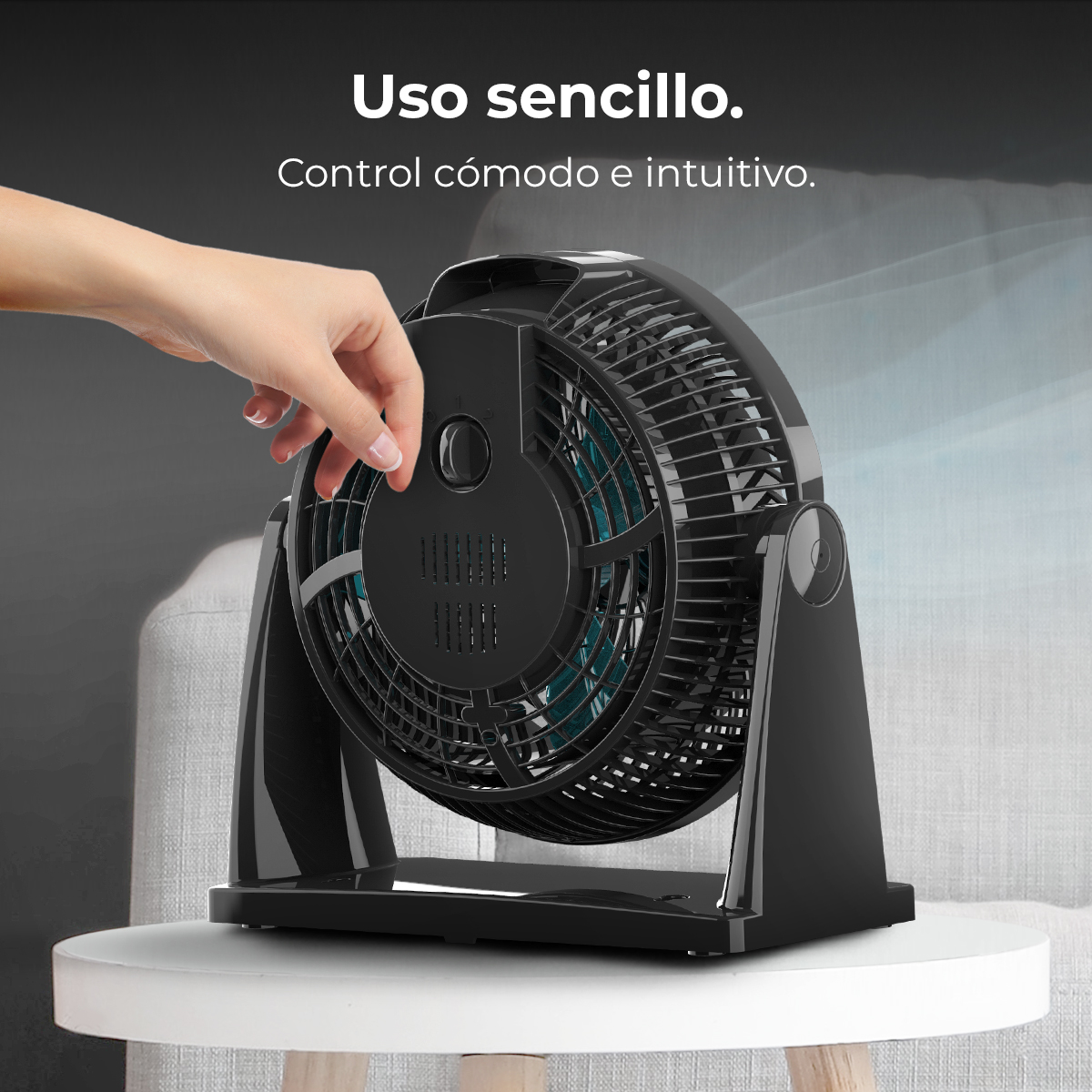 Una mano regola un ventilatore nero su un tavolo bianco. Il ventilatore ha pale blu e si trova davanti a un divano grigio.