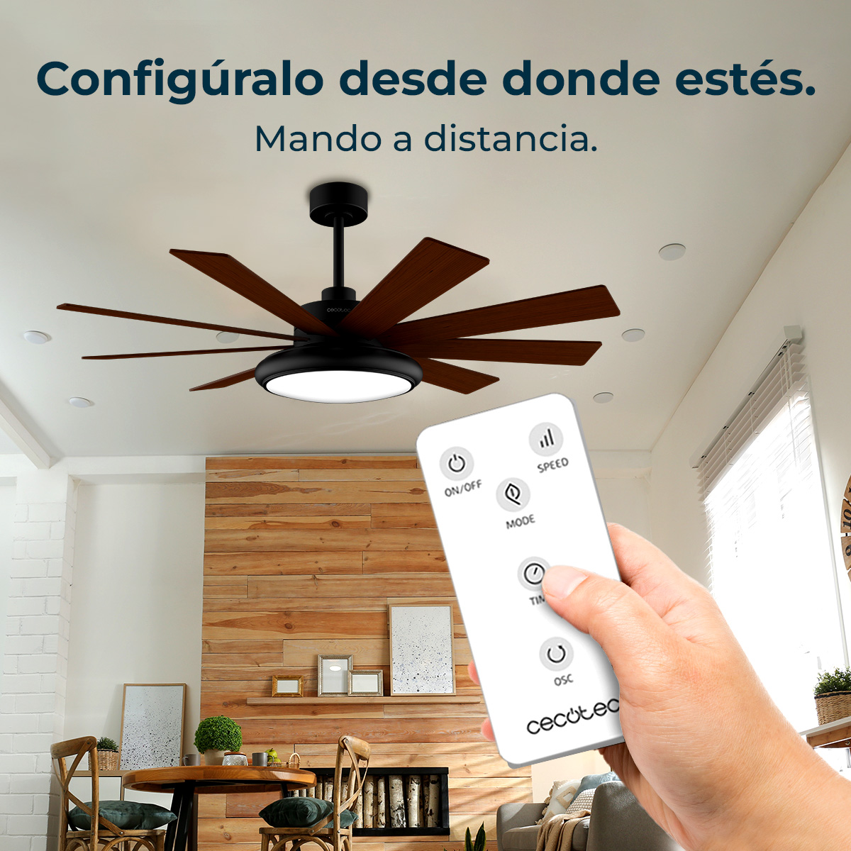 A hand holding a remote controls a ceiling fan. The fan has dark blades and a light. The room has wooden accents and decor.