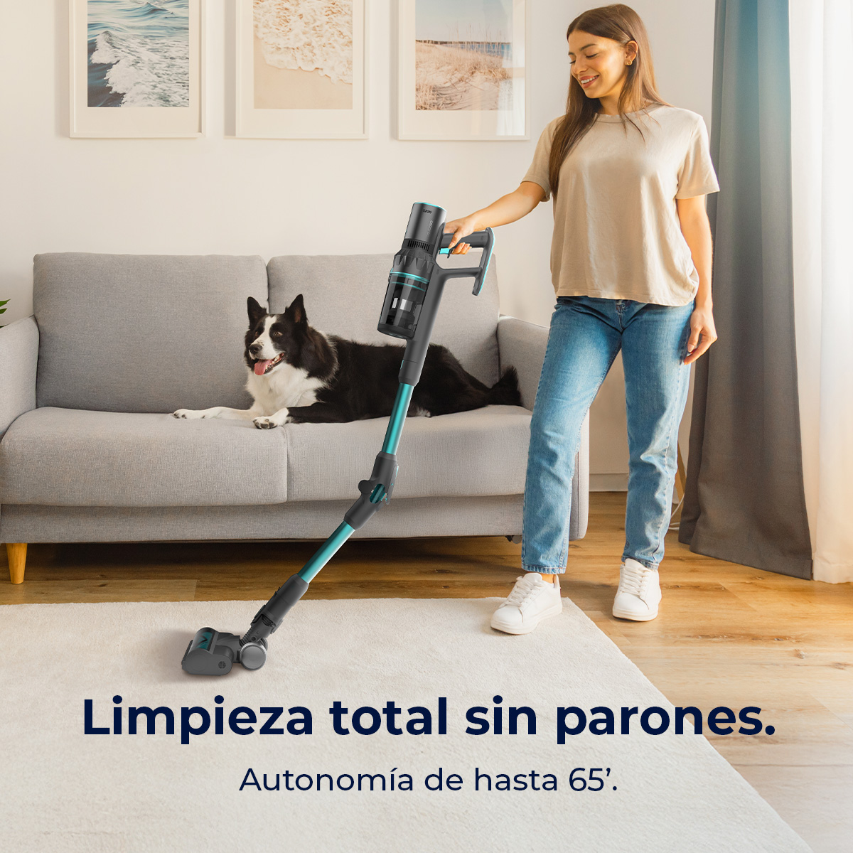 Una mujer aspira una alfombra blanca. Un perro blanco y negro se relaja en un sofá gris. La aspiradora es negra y turquesa.