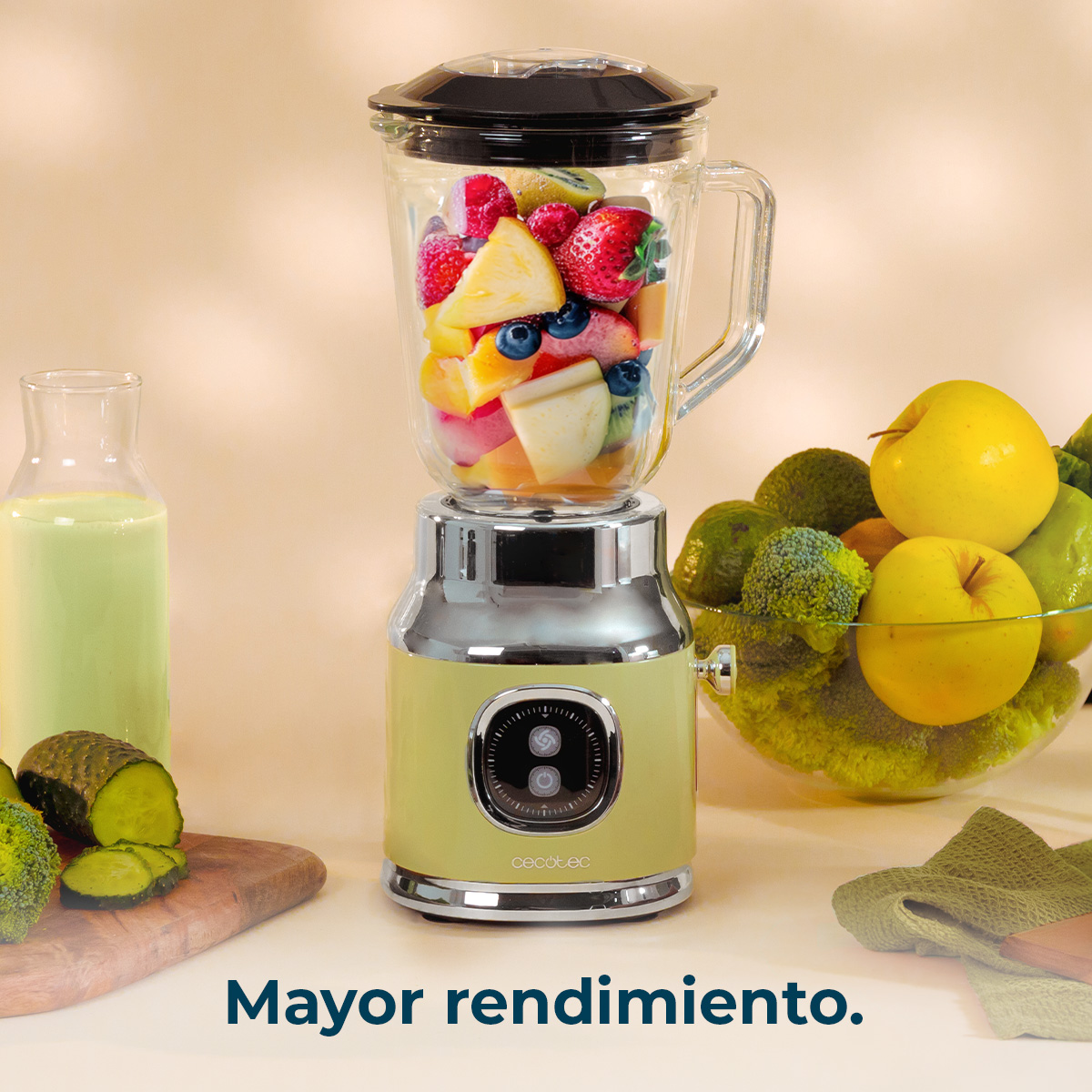 Una licuadora llena de frutas junto a un vaso de líquido verde, con un cuenco de frutas y verduras al fondo.