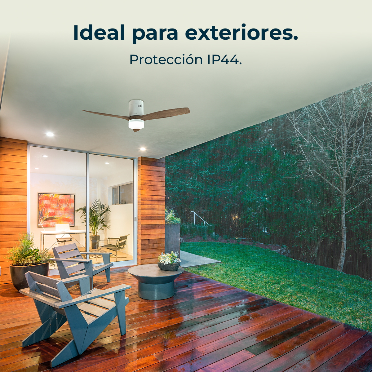 Patio exterior con terraza de madera, sillas, mesa, ventilador de techo. Fondo con césped y árboles, lluvia visible.