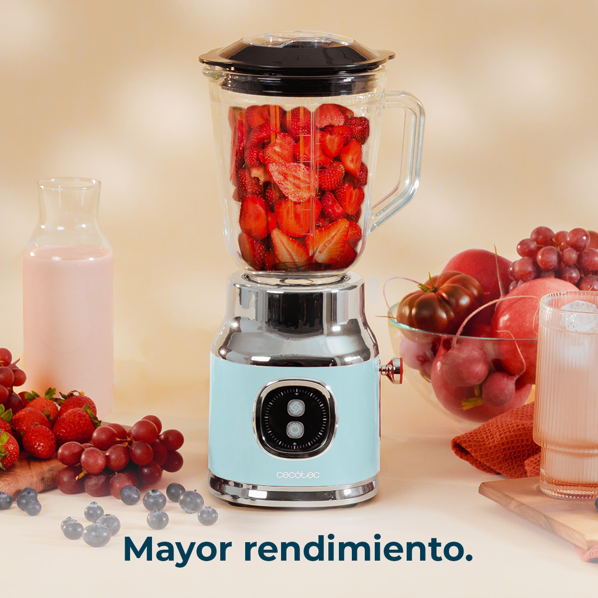 Una batidora llena de fresas está junto a uvas, melocotones y arándanos. A la vista hay un vaso de líquido rosa y una bebida con hielo.