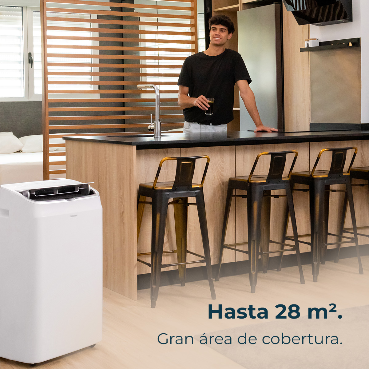 Mężczyzna w kuchni z napojem, klimatyzator na pierwszym planie, stołki barowe. Tekst: 'Hasta 28 m2. Gran área de cobertura.'