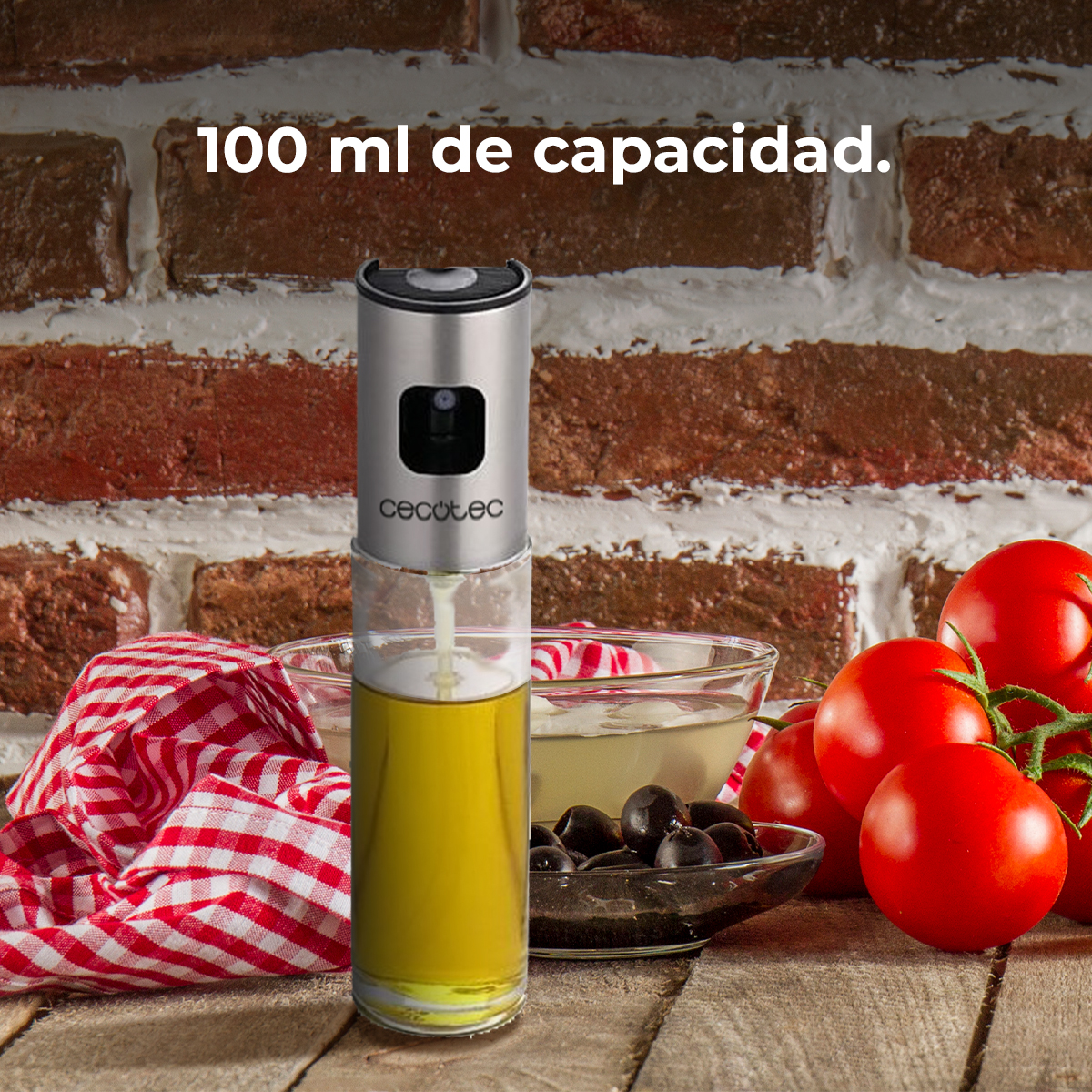 Pulverizador de aceite de oliva con aceite. Tomates, aceitunas y un paño a cuadros rojos están sobre madera, frente a una pared de ladrillos.