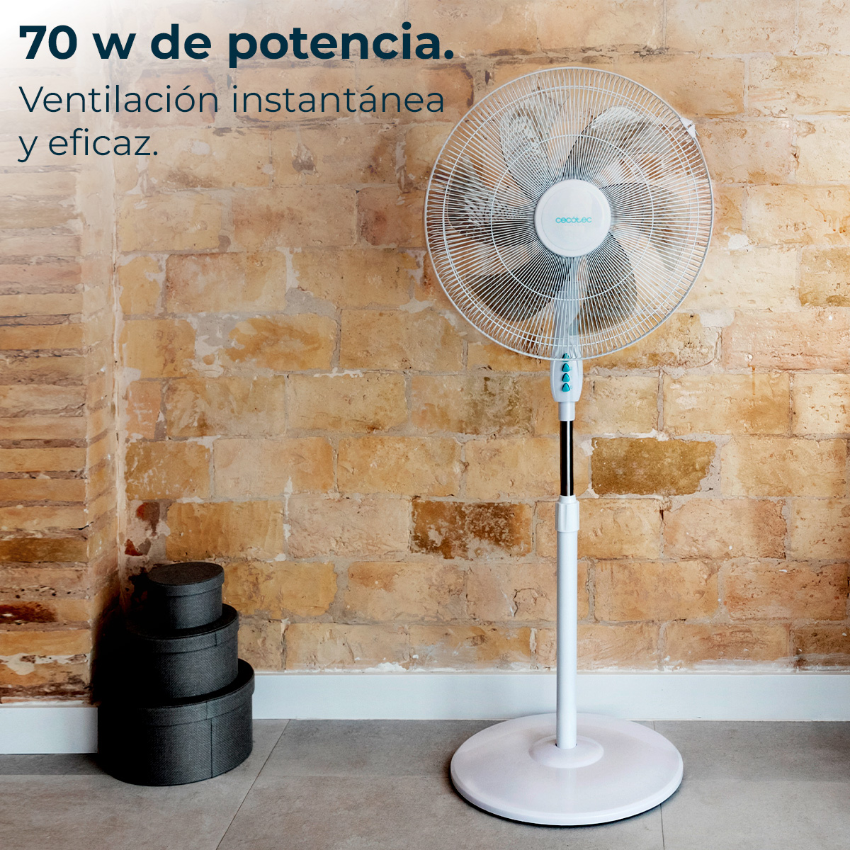 Biały wentylator stojący przed ceglaną ścianą. Trzy czarne okrągłe pudełka po lewej stronie. Tekst nad wentylatorem.