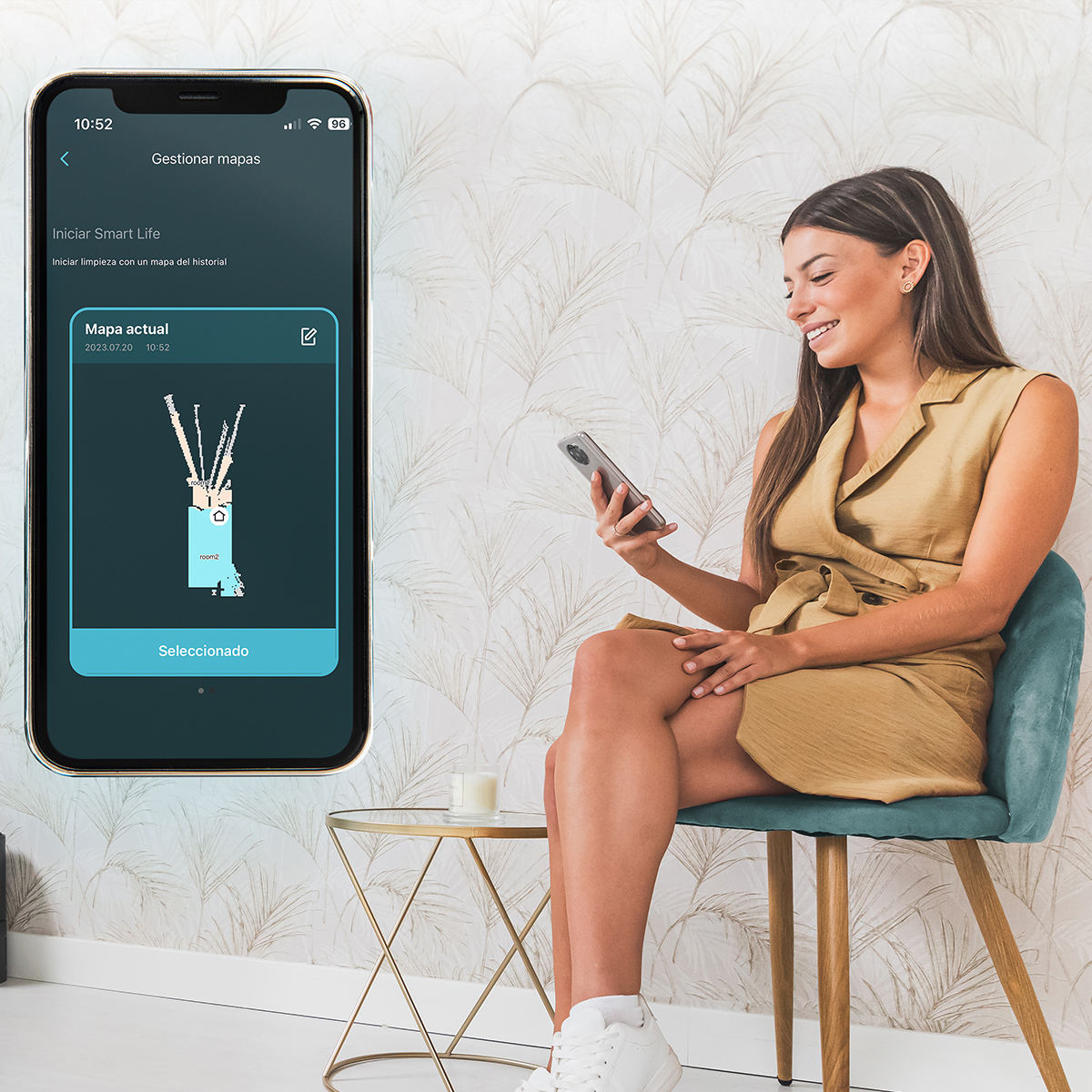 Una mujer sonríe mientras sostiene un teléfono, mirando la pantalla. Un teléfono muestra un mapa, mientras ella está sentada en una silla azul, en una habitación.