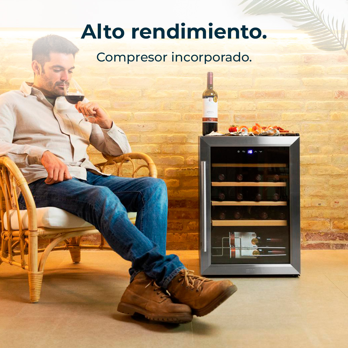 Uomo seduto, che beve vino, vicino a un refrigeratore per vino. Bottiglia e snack sopra. Sfondo muro di mattoni. Indossa jeans e stivali.