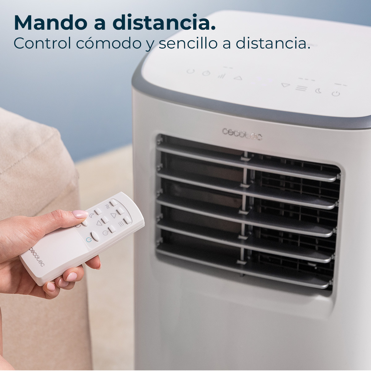 Dłoń trzyma biały pilot w pobliżu białego klimatyzatora. Tłem jest niebieska ściana i beżowa kanapa.