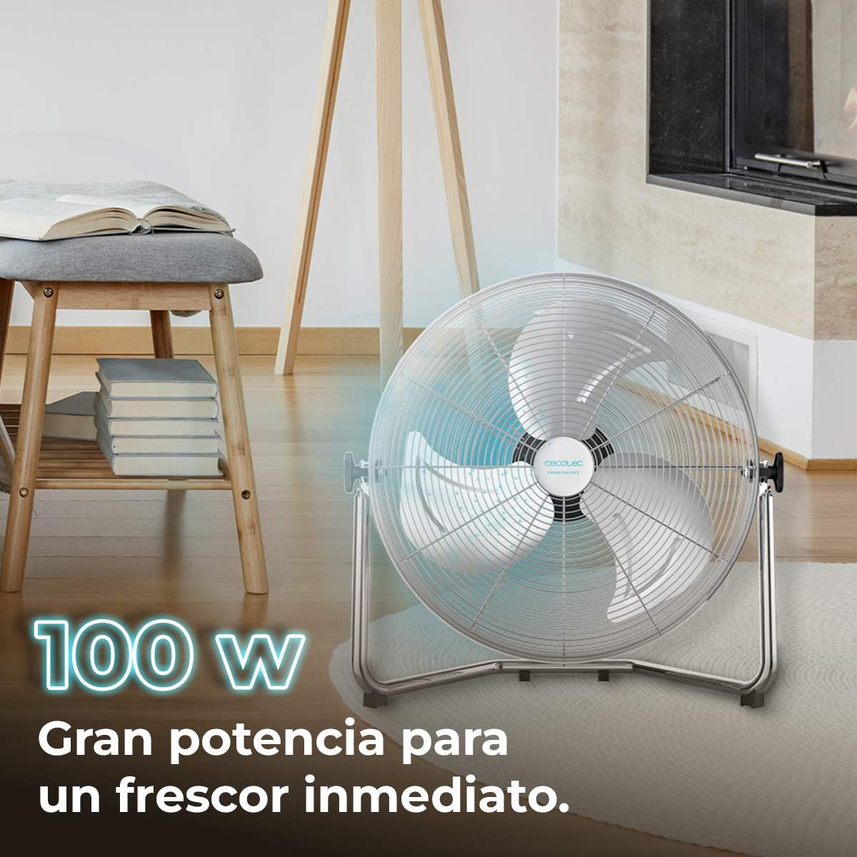 Srebrny wentylator na dywanie. Po lewej stronie drewniany stołek z książkami i lampa podłogowa. Tekst: '100 W' i 'Gran potencia'.