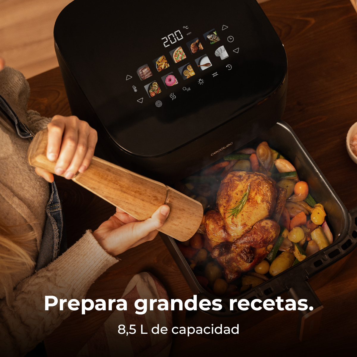 Una freidora de aire con pollo asado y verduras. Una persona sostiene una espátula de madera. La freidora es negra, la comida humea.