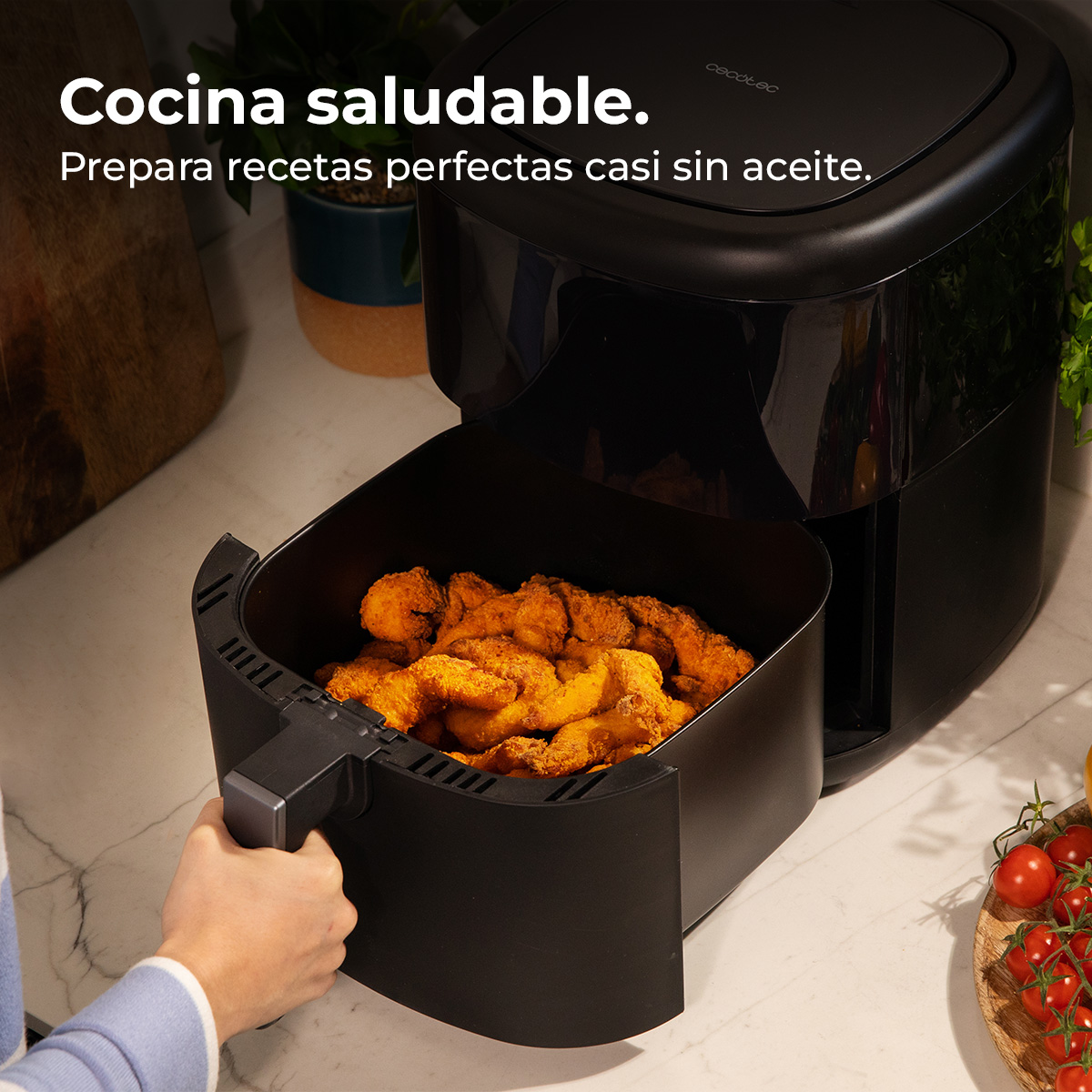 Zwarte airfryer met handvat met gefrituurd voedsel. Tomaten en plant op de achtergrond. Tekst 'gezond koken'.